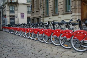 A row of red bicycles with white lettering on the fenders, parked at a docking station on a cobblestone street in front of a building with a purple sign.
