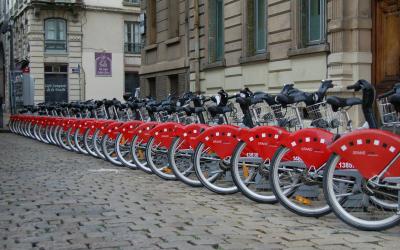 A row of red bicycles with white lettering on the fenders, parked at a docking station on a cobblestone street in front of a building with a purple sign.