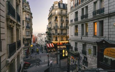 A narrow Parisian street at dusk featuring Haussmann-style buildings, a "METROPOL" hotel sign with red-and-white awnings, and a yellow "Le Marais" directional sign on the right.