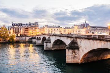 1. A stone arch bridge illuminated at dusk over a river, with classical European-style buildings lining the far bank under a cloudy sky.