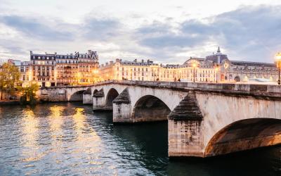 A stone arch bridge illuminated at dusk over a river, with classical European-style buildings lining the far bank under a cloudy sky.