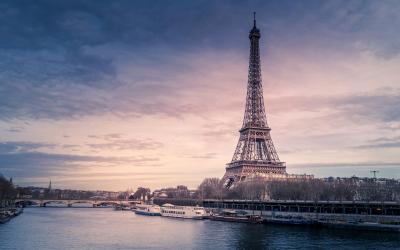 The image shows the Eiffel Tower standing prominently beside the Seine River in Paris, with tour boats on the water and a twilight sky.
