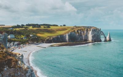 A coastal scene featuring a prominent natural limestone arch (the Porte d'Aval) and a smaller stack (the Porte Amont) at Étretat, Normandy, France, with a sandy beach, cliffs, and green fields above.