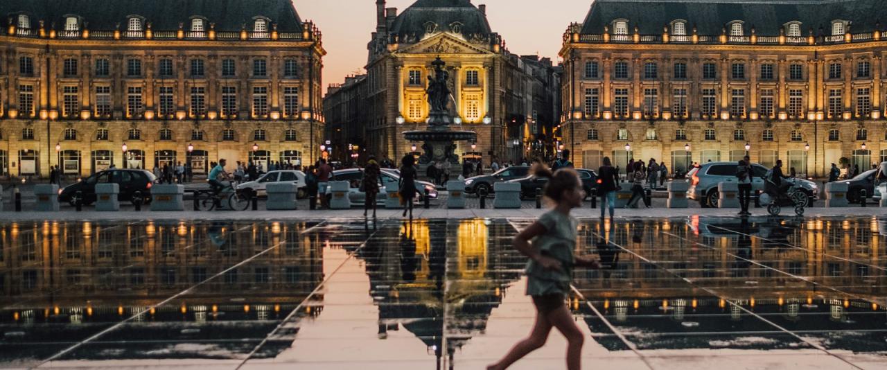 A wide-angle evening shot of Place de la Bourse in Bordeaux, France, featuring the Miroir d'eau (Water Mirror) reflecting the illuminated facades of the 18th-century buildings and the central monument, with a blurred figure of a child running acro
