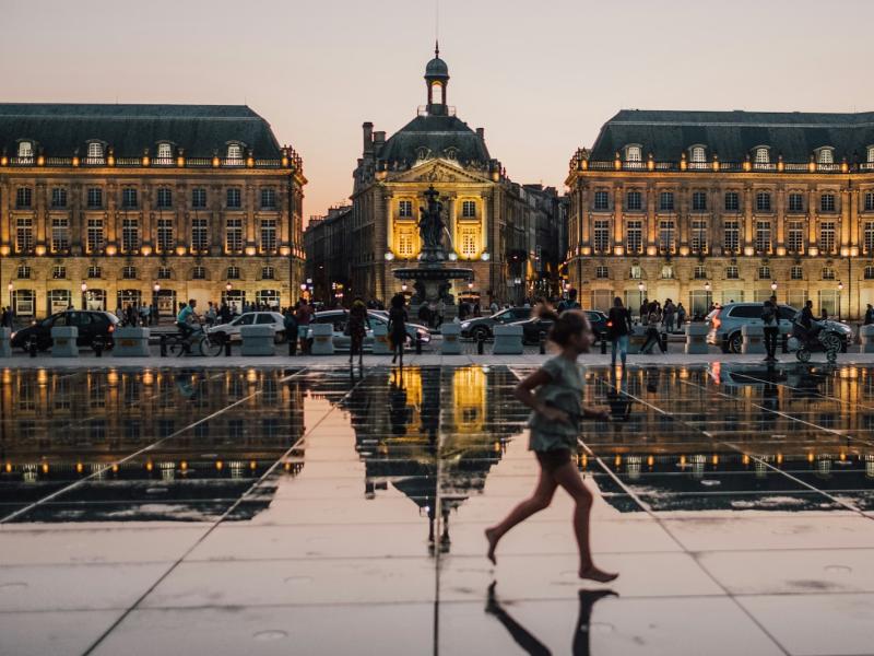 1. A wide-angle evening shot of Place de la Bourse in Bordeaux, France, featuring the Miroir d'eau (Water Mirror) reflecting the illuminated facades of the 18th-century buildings and the central monument, with a blurred figure of a child running acro