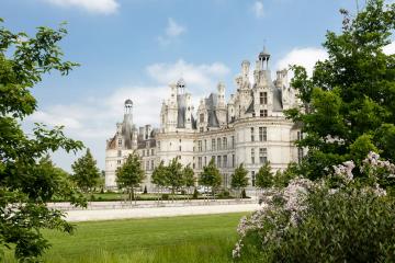 A large, ornate French Renaissance château with multiple towers, chimneys, and a central dome, viewed from across a manicured lawn with trees framing the foreground under a blue sky.