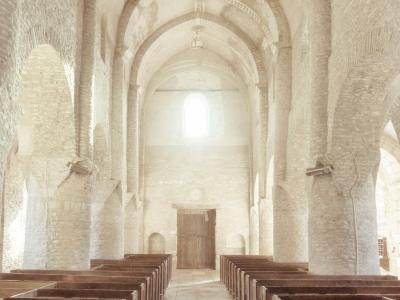 1. The image shows the interior nave of a stone church with massive rounded arches, thick piers, and a barrel-vaulted ceiling, illuminated by bright light from a high window at the far end; wooden pews line the aisle.