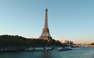 The image shows the Eiffel Tower standing prominently beside the Seine River in Paris, with tour boats on the water and a twilight sky.