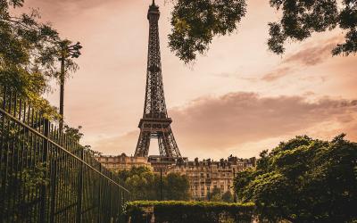 The image shows the Eiffel Tower in Paris, France, viewed through a frame of green trees and foliage with a metal fence in the foreground under a warm-toned sky.