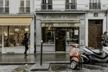 A red motor scooter is parked on a wet Parisian sidewalk in front of storefronts, including one with a sign reading "Le Carillon d'Olivier" and another with an orange awning.