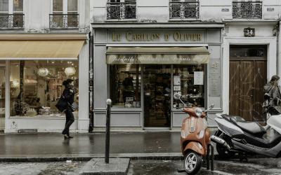 A red motor scooter is parked on a wet Parisian sidewalk in front of storefronts, including one with a sign reading "Le Carillon d'Olivier" and another with an orange awning.