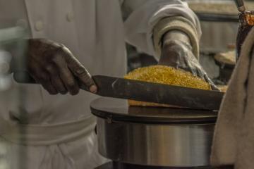 1. A close-up shot of a chef's hands using a long, serrated knife to slice a golden-brown baked good, possibly a cake or bread, on a metal stand in a professional kitchen setting.