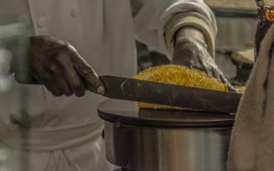 A close-up shot of a chef's hands using a long, serrated knife to slice a golden-brown baked good, possibly a cake or bread, on a metal stand in a professional kitchen setting.