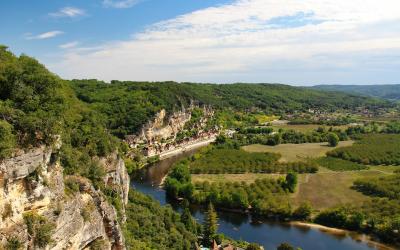 A high-angle landscape view of a winding river flowing through a valley with steep limestone cliffs on the left, dense green forests, and a small town nestled along the riverbank under a blue sky with scattered clouds.