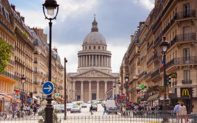 A street scene in Paris, France, looking down Rue de l'Odéon towards the Panthéon, with classic Haussmannian architecture, a McDonald's on the right, and a blue one-way traffic sign on the left.