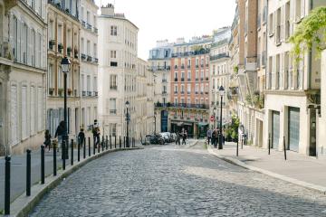 1. A sunlit, curving cobblestone street lined with multi-story Haussmann-style buildings featuring wrought-iron balconies and shuttered windows, with pedestrians and parked cars visible in the distance.