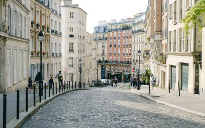 A sunlit, curving cobblestone street lined with multi-story Haussmann-style buildings featuring wrought-iron balconies and shuttered windows, with pedestrians and parked cars visible in the distance.