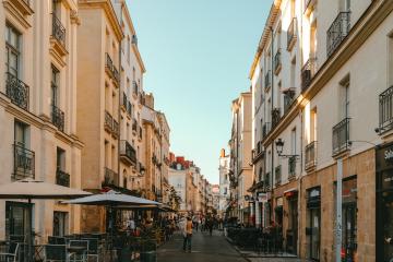 1. A narrow, sunlit pedestrian street lined with multi-story buildings featuring wrought-iron balconies and ground-floor cafes or shops, including one with a visible “Sélect” sign.