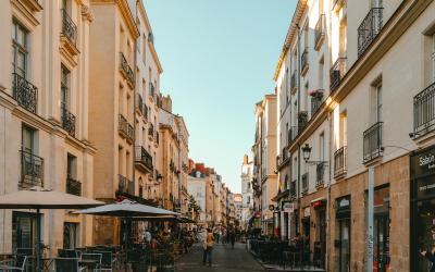 A narrow, sunlit pedestrian street lined with multi-story buildings featuring wrought-iron balconies and ground-floor cafes or shops, including one with a visible “Sélect” sign.
