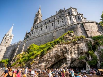 1. The image shows the Basilica of the Immaculate Conception (Upper Basilica) perched atop a rocky cliff in Lourdes, France, with the Grotto of Massabielle visible at its base and crowds of visitors below.