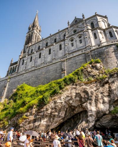1. The image shows the Basilica of the Immaculate Conception (Upper Basilica) perched atop a rocky cliff in Lourdes, France, with the Grotto of Massabielle visible at its base and crowds of visitors below.