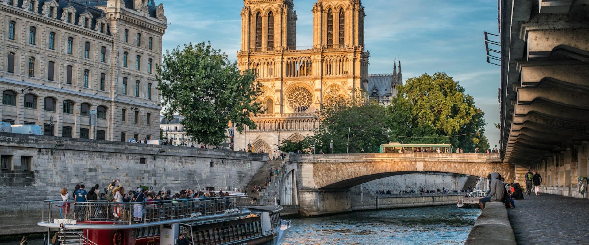 A sightseeing boat on the Seine River with Notre-Dame Cathedral and adjacent Parisian buildings visible in the background under a partly cloudy sky.