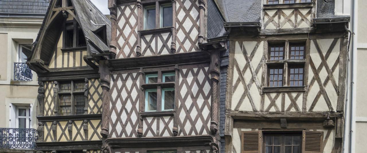 The image shows a row of historic half-timbered houses with distinctive diamond-patterned wooden framing, multi-paned windows, and steep gabled roofs under an overcast sky; no visible signage or text is present.