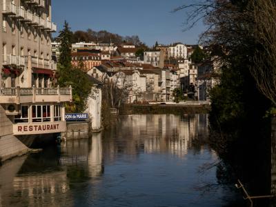 A river flowing through a town with multi-story buildings on the left bank, including a restaurant with a sign reading "RESTAURANT" and another sign that appears to say "GAVE DE PAU", under a clear blue sky.