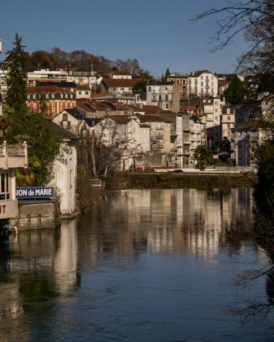 A river flowing through a town with multi-story buildings on the left bank, including a restaurant with a sign reading "RESTAURANT" and another sign that appears to say "GAVE DE PAU", under a clear blue sky.