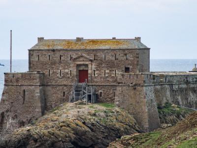 1. A stone coastal fortification with a central rectangular building featuring a red door, situated on a rocky promontory overlooking the sea, with small islands visible in the background.