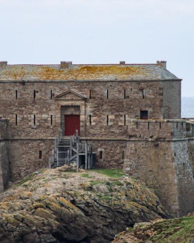 1. A stone coastal fortification with a central rectangular building featuring a red door, situated on a rocky promontory overlooking the sea, with small islands visible in the background.