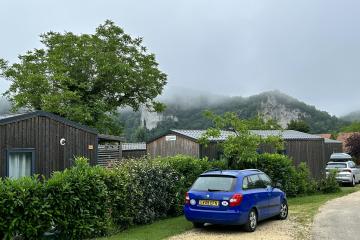 1. A gravel driveway with a blue hatchback and other cars parked near modern wooden cabins or gîtes, bordered by hedges, with misty mountains in the background.