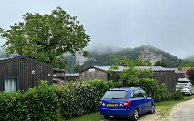A gravel driveway with a blue hatchback and other cars parked near modern wooden cabins or gîtes, bordered by hedges, with misty mountains in the background.