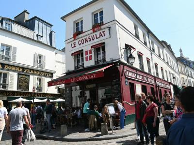 1. The image shows a street corner in a French town featuring a prominent white building with a red awning labeled "LE CONSULAT" and an adjacent shop "LA BORNE FRANCHETTE," with pedestrians walking on the sidewalk under a clear sky.