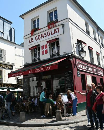 1. The image shows a street corner in a French town featuring a prominent white building with a red awning labeled "LE CONSULAT" and an adjacent shop "LA BORNE FRANCHETTE," with pedestrians walking on the sidewalk under a clear sky.