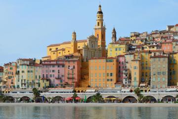 A colorful hillside town with densely packed pastel buildings and a prominent church tower overlooking a waterfront promenade and calm sea.