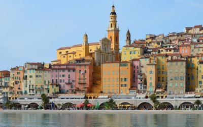 A colorful hillside town with densely packed pastel buildings and a prominent church tower overlooking a waterfront promenade and calm sea.