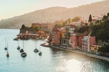 1. A scenic coastal view of Villefranche-sur-Mer, France, showing its harbor with moored sailboats, colorful buildings along the waterfront, and a hillside backdrop under warm sunlight.