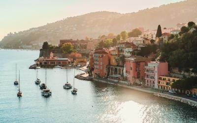 A scenic coastal view of Villefranche-sur-Mer, France, showing its harbor with moored sailboats, colorful buildings along the waterfront, and a hillside backdrop under warm sunlight.
