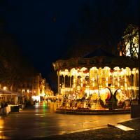 1. A brightly lit carousel stands in a paved city square at night, surrounded by trees, market stalls with white canopies, and classical European-style buildings; no visible signage identifies the location.