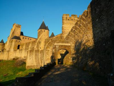 The image shows a sunlit medieval stone fortress with high defensive walls, crenellated towers with conical roofs, and an arched gateway, set against a clear blue sky with a grassy slope in the foreground.
