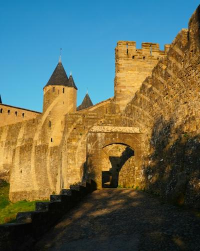 The image shows a sunlit medieval stone fortress with high defensive walls, crenellated towers with conical roofs, and an arched gateway, set against a clear blue sky with a grassy slope in the foreground.