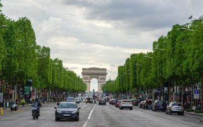 A wide, tree-lined avenue in Paris with traffic including cars and motorcycles, leading toward the Arc de Triomphe in the distance under an overcast sky.