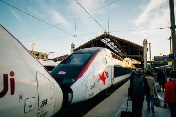 1. The image shows two high-speed trains at a station platform, with passengers walking nearby; the train on the right displays a red logo resembling the TGV branding and has visible numbering (e.g., “266”), while the left train shows partial letteri