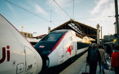 The image shows two high-speed trains at a station platform, with passengers walking nearby; the train on the right displays a red logo resembling the TGV branding and has visible numbering (e.g., “266”), while the left train shows partial letteri