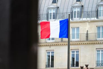 1. A French tricolor flag on a pole in front of a multi-story building with mansard roof and dormer windows, viewed through a dark foreground obstruction.