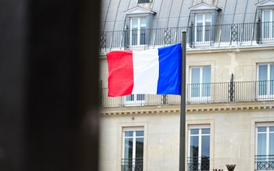 A French tricolor flag on a pole in front of a multi-story building with mansard roof and dormer windows, viewed through a dark foreground obstruction.