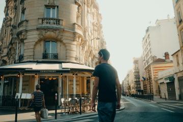 1. A street corner in a European city featuring a corner café with a striped awning, a man walking away from the camera, and classic multi-story buildings with balconies and large windows.