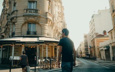 A street corner in a European city featuring a corner café with a striped awning, a man walking away from the camera, and classic multi-story buildings with balconies and large windows.