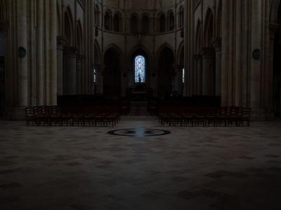 1. The image shows the dimly lit interior nave of a large stone church, featuring tall columns, arched windows, and rows of wooden chairs facing an altar area with a stained-glass window at the far end.